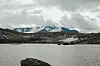 Fjellvann med steiner i forgrunnen og snødekte fjell under skyet himmel.